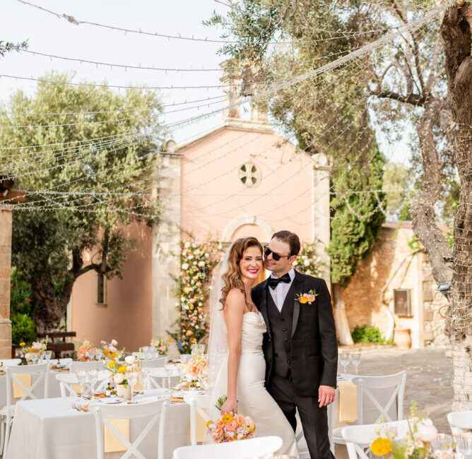 Bride and groom posing at outdoor wedding reception with white tables, yellow flowers, and historic stone chapel