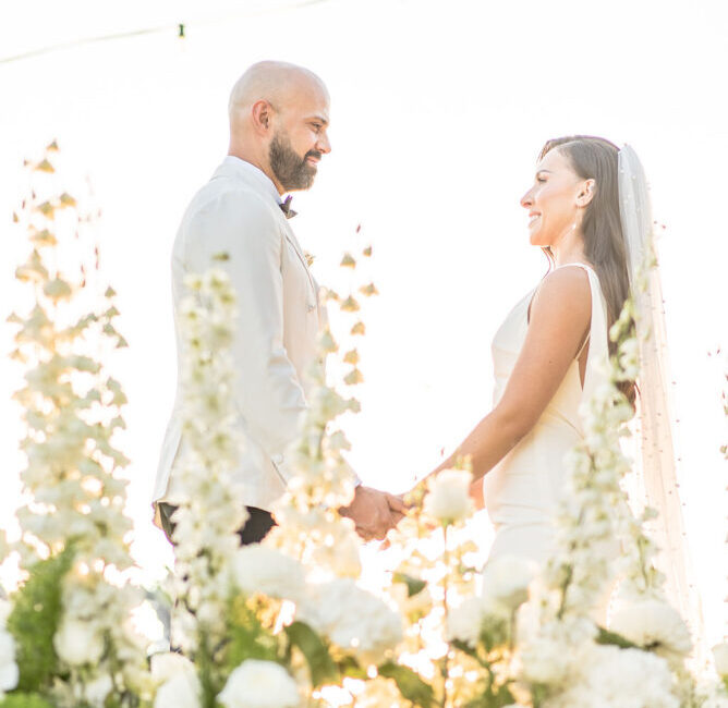 Bride and groom holding hands during wedding ceremony surrounded by white flowers