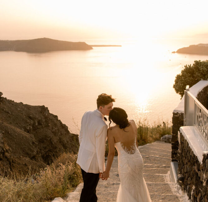 Bride and groom kissing at sunset on cliffside terrace overlooking calm Mediterranean bay with islands