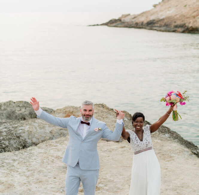 Newlyweds celebrating on beach with rocky cliffs, bride holding pink bouquet, groom in light blue suit