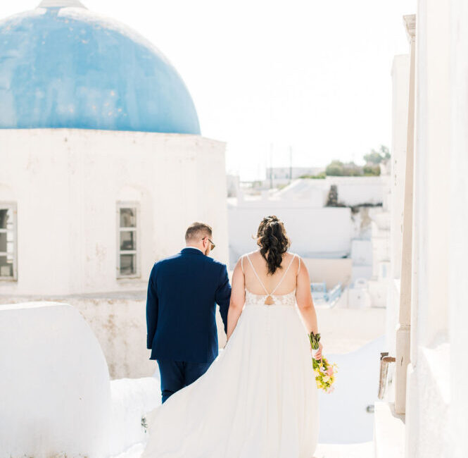 Bride and groom walking together through white-washed streets with blue dome church in Santorini