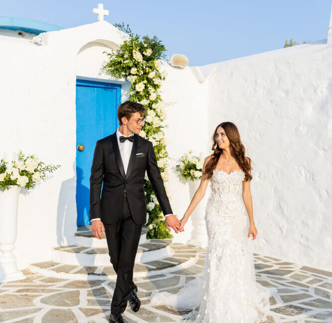 Bride and groom holding hands walking past white chapel with blue door in Mediterranean setting