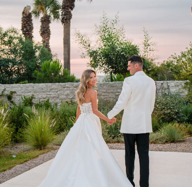 Bride and groom holding hands at sunset in elegant garden with palm trees and stone wall backdrop