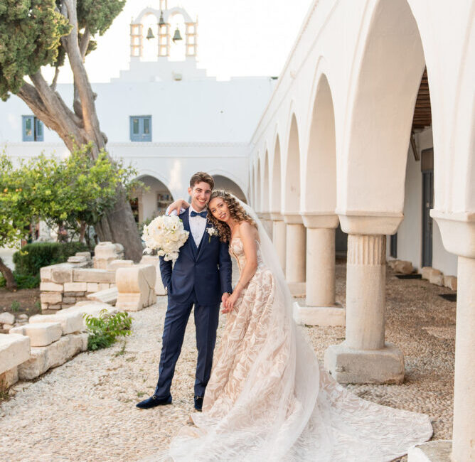 Bride and groom posing in Mediterranean courtyard with white arches and church dome backdrop