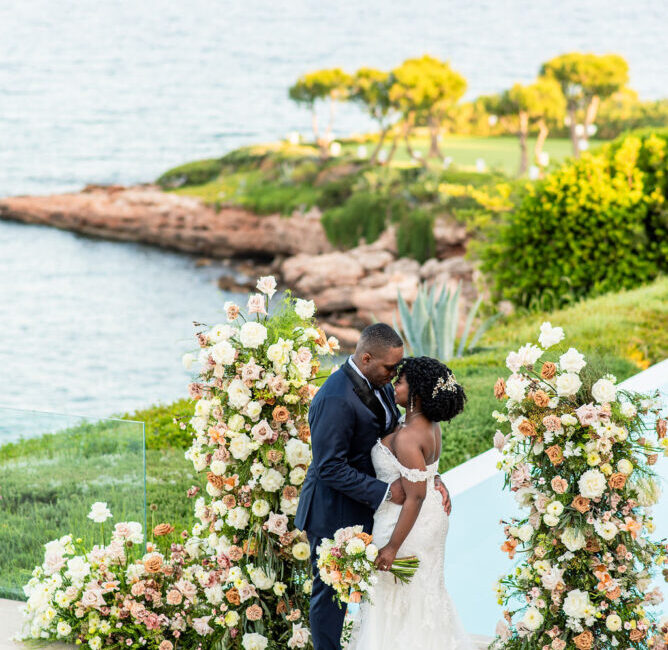 Bride and groom kissing at seaside wedding ceremony with white and peach floral arch